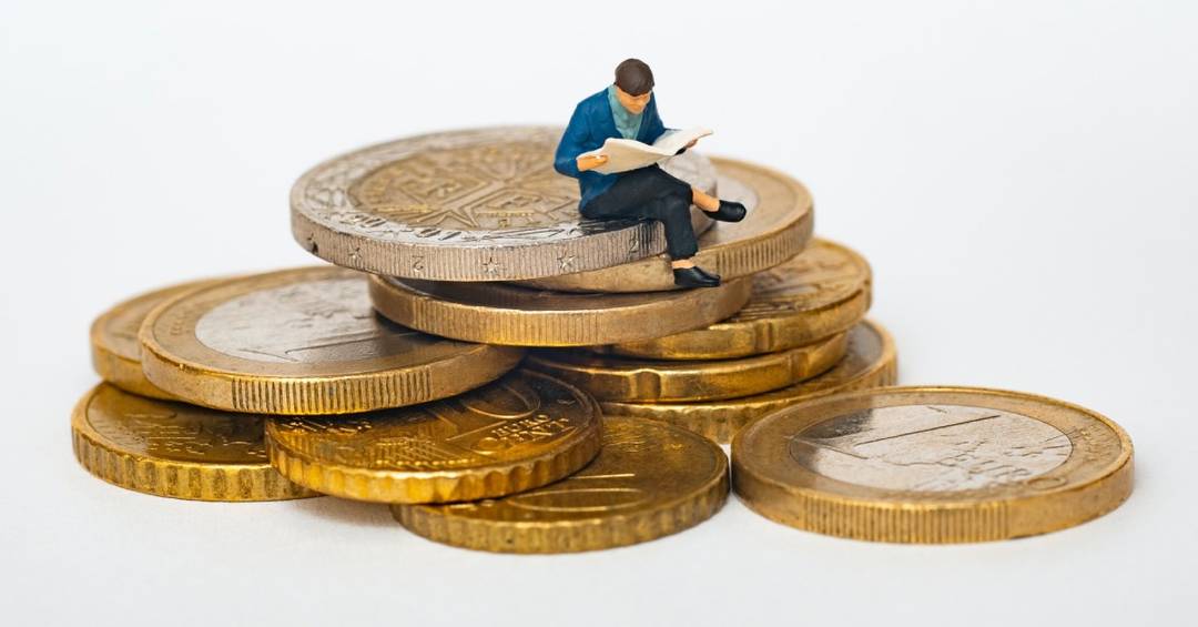 Eine kleine Figur sitzt auf einem Stapel Euro-Münzen und liest in einem Buch