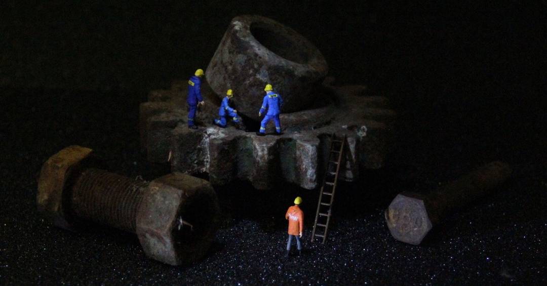 Vier kleine Miniaturfiguren im blauen und orangenen THW-Outfit klettern gemeinsam mit einer Leiter auf ein großes verrostetes Zahnrad aus Metall