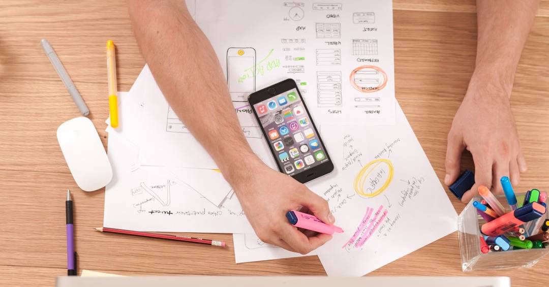 Blick auf einen Schreibtisch, auf dem zwei Arme mit bunt markierten Zetteln, einem Smartphone und einem Computer Werbung für einen Sportverein erstellen.