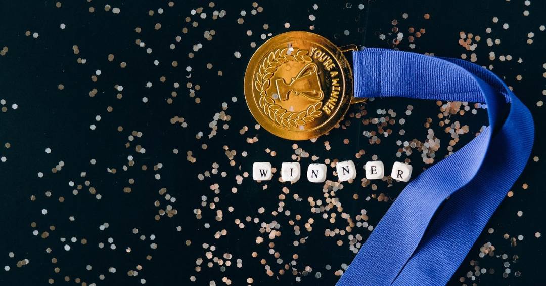 Eine goldene Medaille an einem blauen Band vor schwarzem Hintergrund, daneben silberner Glitter und der Schriftzug "WINNER"