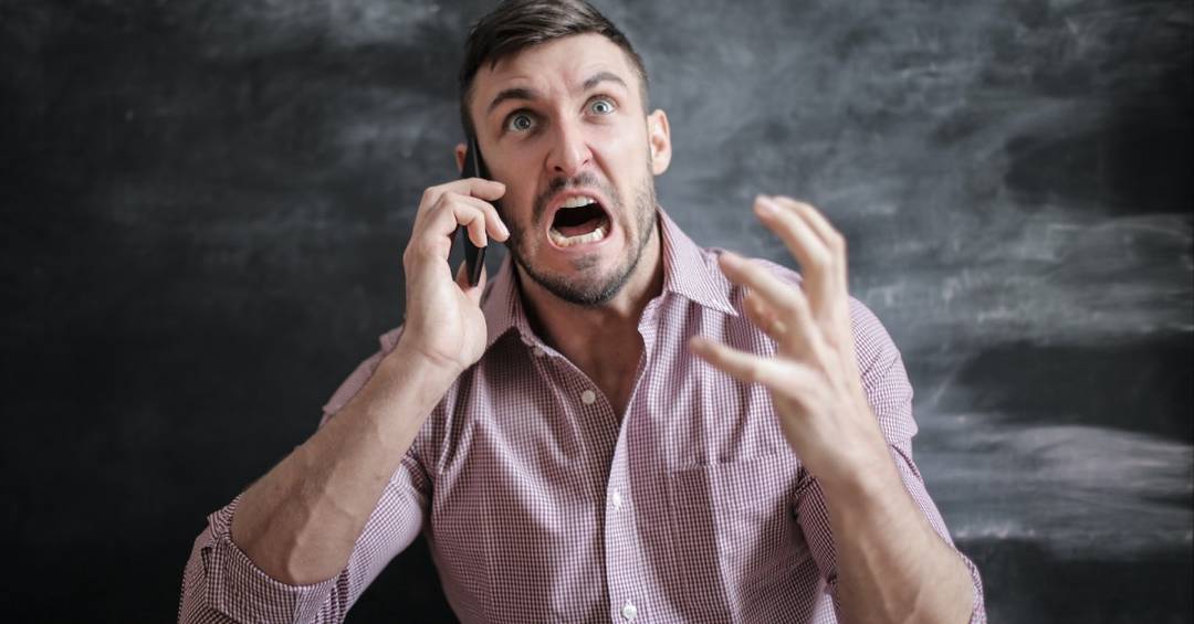 Ein junger Mann im karierten Hemd telefoniert und schaut schockiert nach oben, dazu gestikuliert er wild.